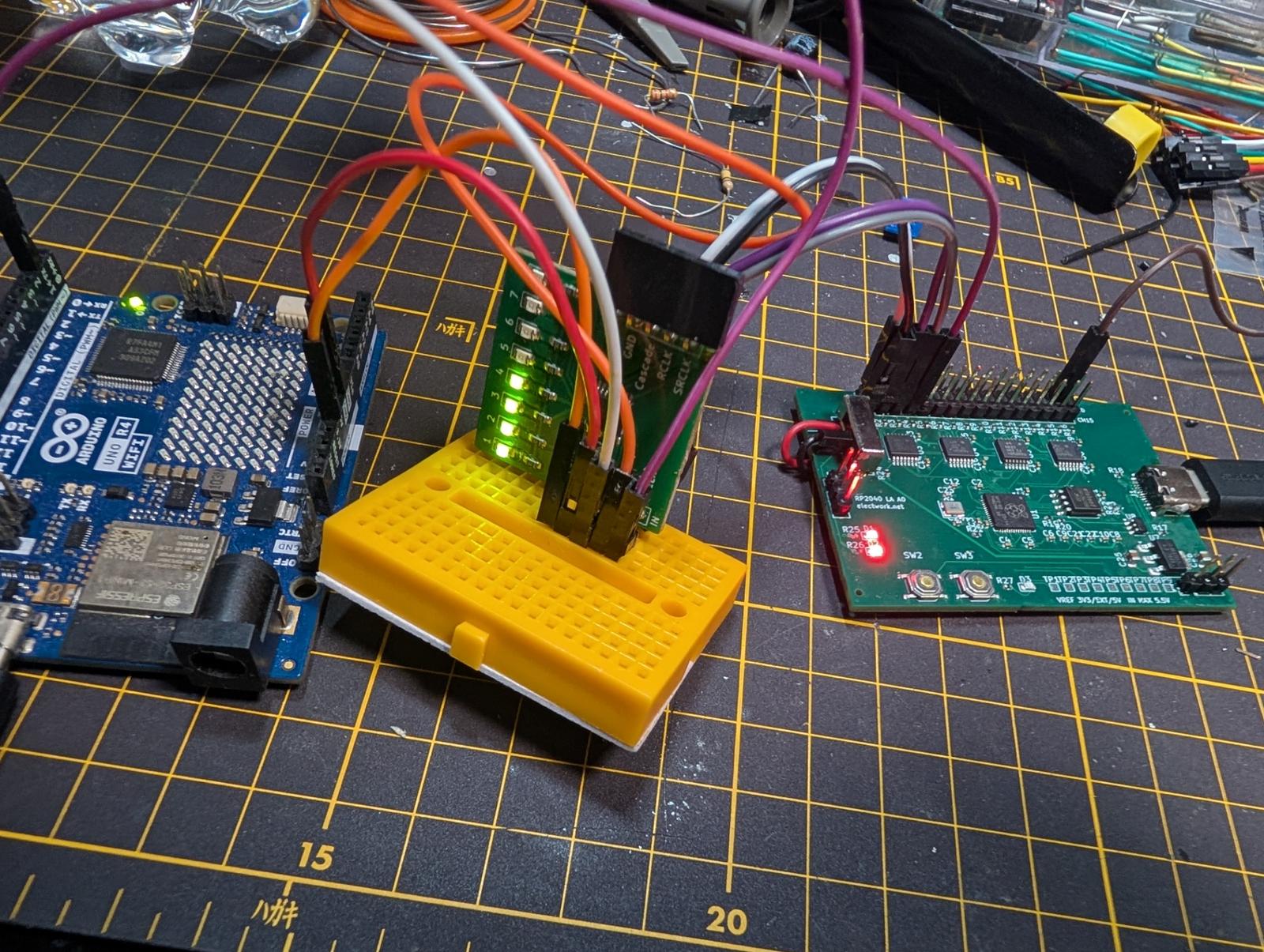 机の上の写真。左にArduino UNO R4 WiFi、中央にミニブレッドボードに挿した74HC595のLED基板で1から4のLEDが緑に点灯、右に自作のRP2040ロジアナ基板